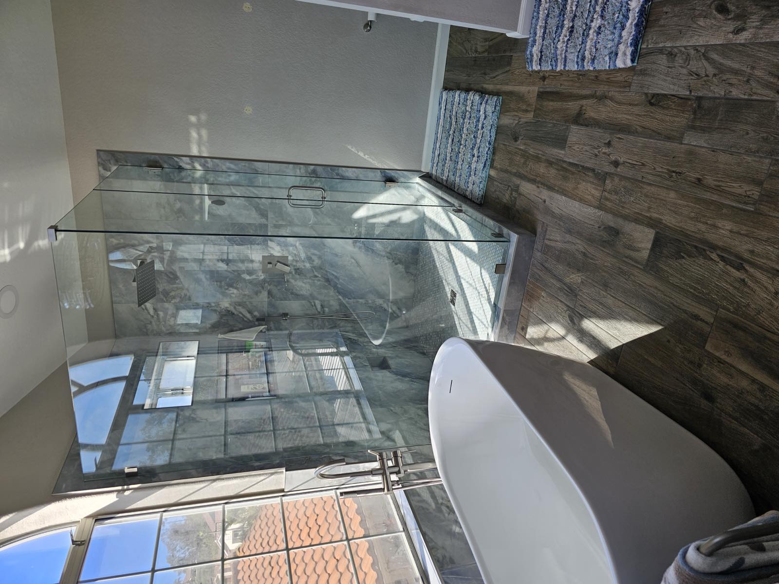 Blue marble walk-in glass shower beside a freestanding soaking tub with wood-look tile floors
