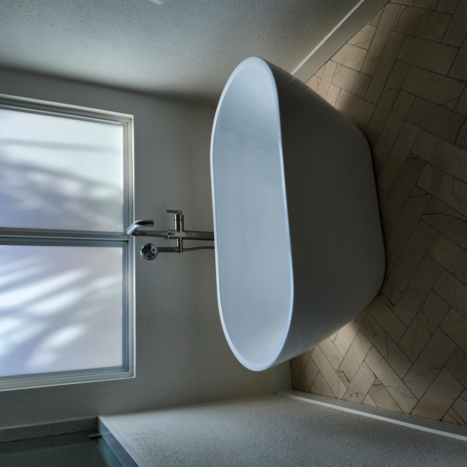 Freestanding soaking tub on a herringbone marble tile floor beneath a frosted window