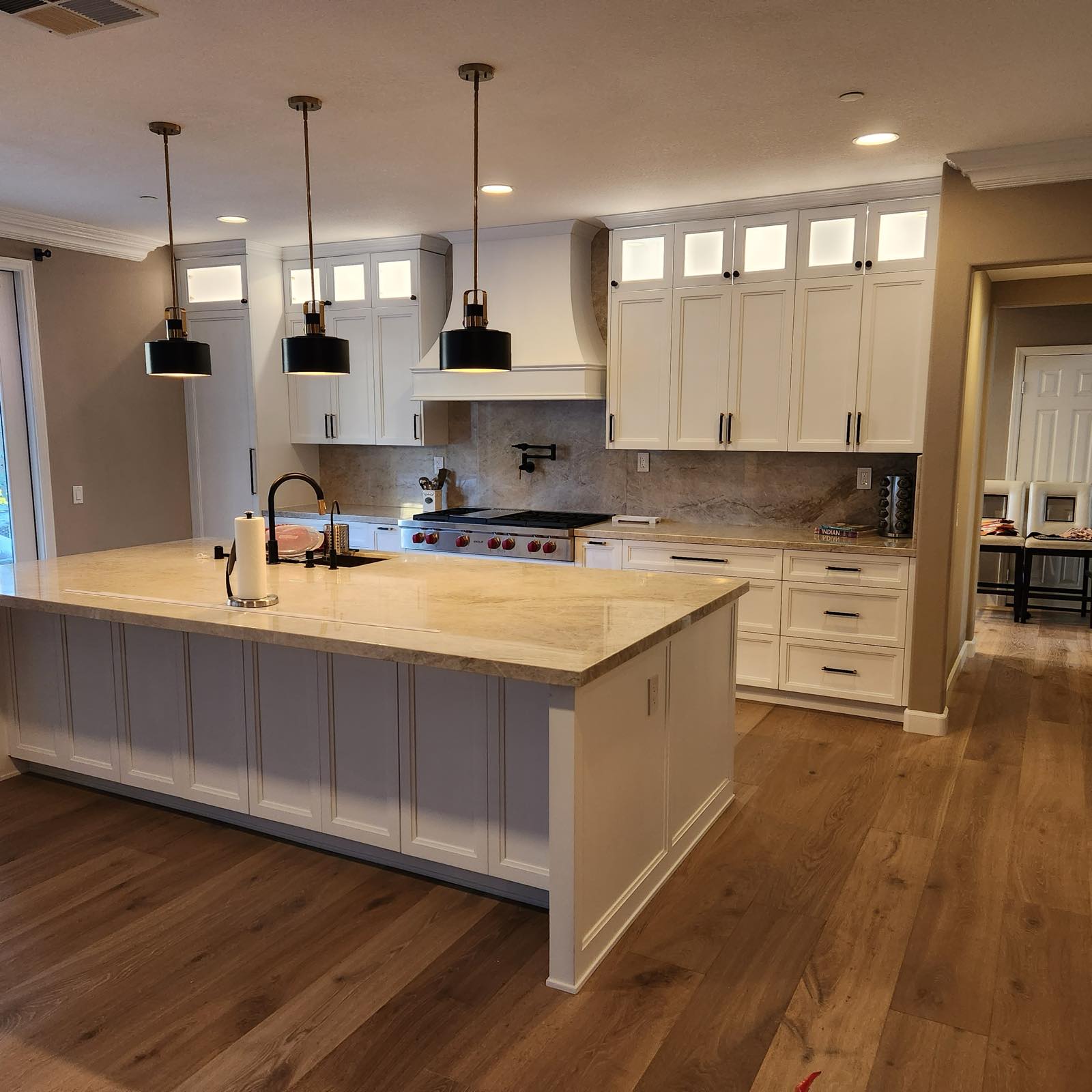 White shaker kitchen with a large island, custom range hood, stone backsplash, and wide-plank wood floors